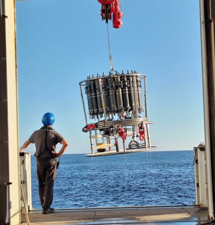 Per Kran wird ein Probensammler von einem Forschungsschiff ins Meer gelassen, er wird Wasserproben in verschiedenen Tiefen sammeln. Bild: Rieke Schäfer/PTB
